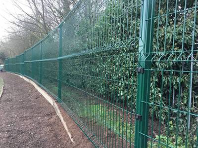 A dark green PVC coated welded wire 3D security fence with 3 curves is installed between the road and trees.