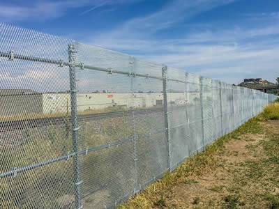 A high expanded metal security fence with three rails is installed between a factory and farm land.