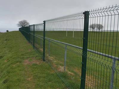 A dark green PVC coated welded 3D security fence with 3 curves is installed in a piece of farm land.