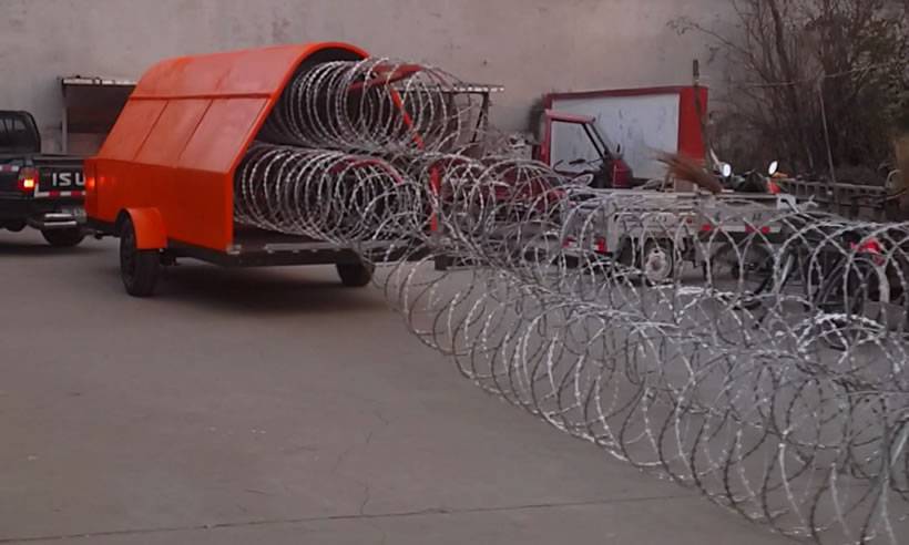 An orange razor wire trailer is stretching concertina razor wire. And the wire has been stretched for a long distance.